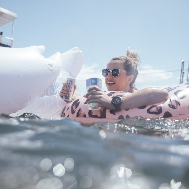 A woman in sunglasses is lounging on an inflatable float in the water, holding a canned drink in each hand. Other inflatables and parts of a boat are visible in the background, hinting at a lively catamaran party Sydney.