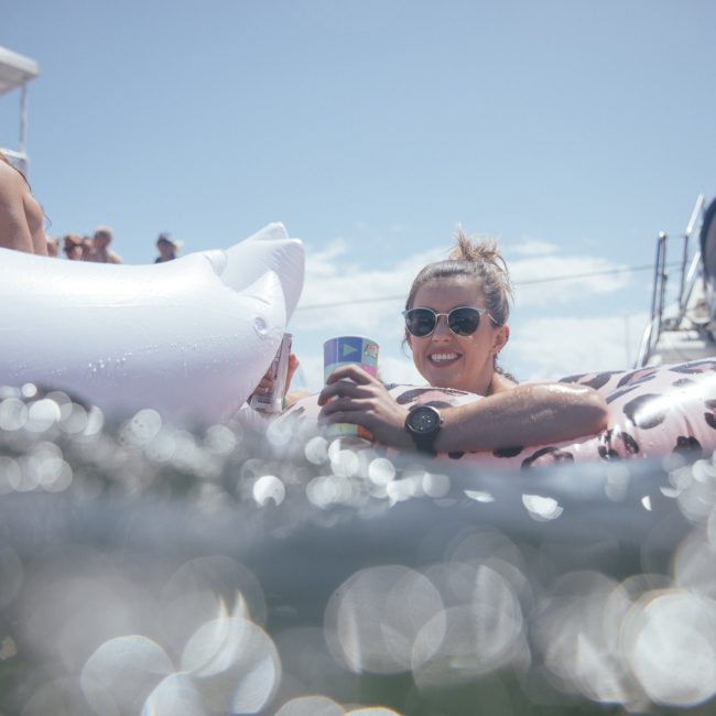 A woman in sunglasses floats on an inflatable in the water, holding a drink. A boat is in the background with people enjoying a Sydney boat party hire. The day appears sunny and bright.