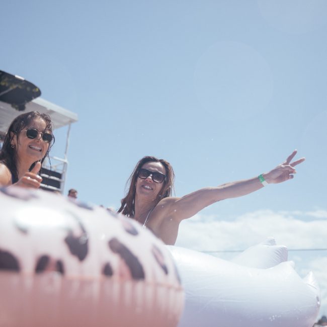 Two women in sunglasses enjoy a sunny day in a swimming pool, one making a peace sign. Inflatable pool toys are visible in the foreground against a clear blue sky, reminiscent of a luxurious private yacht charter on Sydney Harbour.