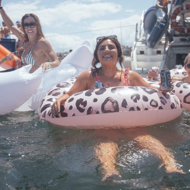 Four women are floating in the water on inflatable pool floats, smiling and holding drinks, with more people and boats visible in the background, enjoying what seems to be a fun Sydney boat party hire.