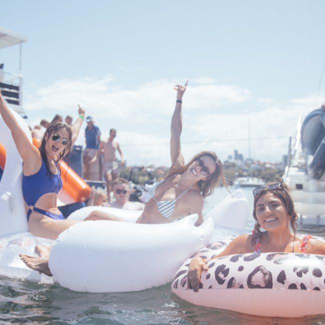 A group of people enjoying a sunny day on the water, sitting on inflatable floats and smiling. Several boats, including luxury yacht hire Sydney options, are visible in the background.