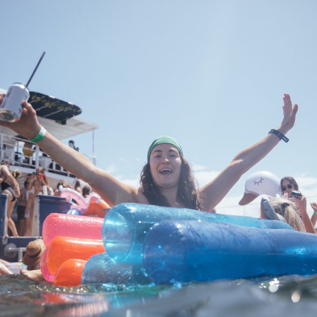 A person with raised arms, wearing a green headband, sits on vibrant inflatable pool floats in the water near a docked boat filled with people enjoying a private yacht charter in Sydney Harbour.