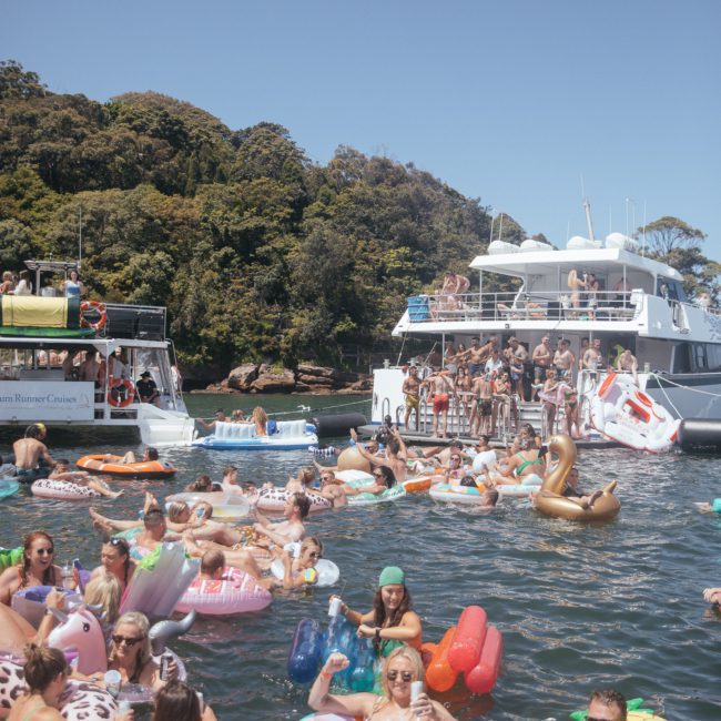 A crowded scene of numerous people enjoying a sunny day on various colorful inflatable floats in the water, with two large boats nearby and a wooded shoreline in the background. This vibrant gathering includes a Catamaran party Sydney, adding to the lively atmosphere.
