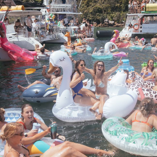 People enjoy a sunny day on a lake, lounging on various inflatable floats resembling swans, dolphins, and other animals. In the background, boats glimmer in the sunlight like those from a luxury yacht hire in Sydney, surrounded by lush greenery.
