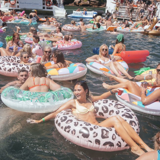 People enjoying a summer day in a lake, floating on colorful inflatable tubes and rafts, with private yacht charters in the background.
