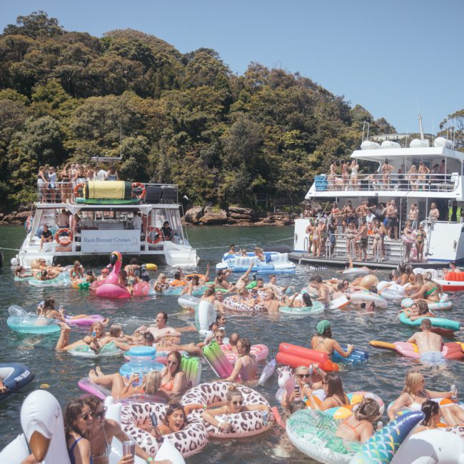 A large group of people enjoys a day on the water, swimming and floating on colorful inflatables. Two boats, anchored nearby, host additional guests. Trees line the background, creating a perfect setting for a Sydney boat party hire or a private yacht charter around Sydney Harbour.