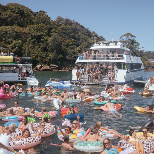 A large group of people are floating in a river on inflatable rafts and pool toys near a private yacht charter in a forested area on a sunny day.