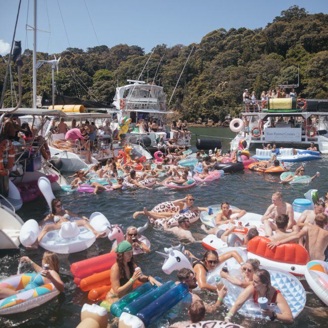 A crowded water scene with numerous people on inflatable floats, surrounded by boats, including a luxury yacht hire Sydney, under a clear sky with lush greenery in the background.