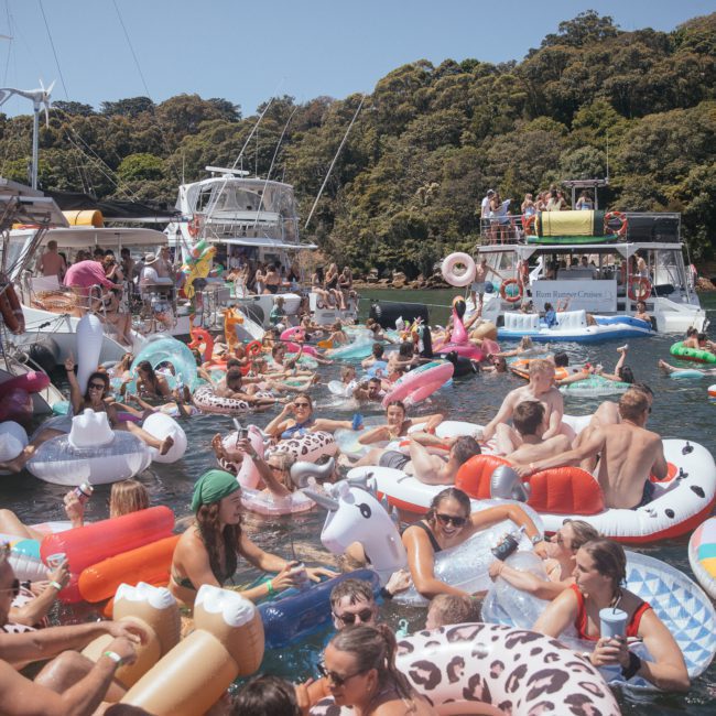 A large group of people enjoying a sunny day on a lake, floating on colorful inflatables, with several boats anchored nearby and dense greenery in the background. Consider a Private yacht charter Sydney Harbour for your next adventure.