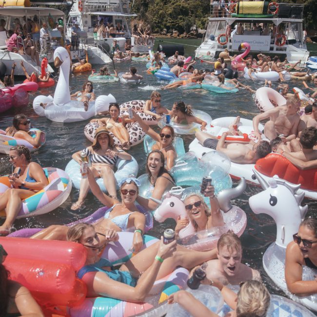 A vibrant pool party with numerous people on colorful inflatable floats shaped like animals and other objects, enjoying themselves. Several boats, including a stylish DJ boat hire Sydney vessel, are visible in the background.