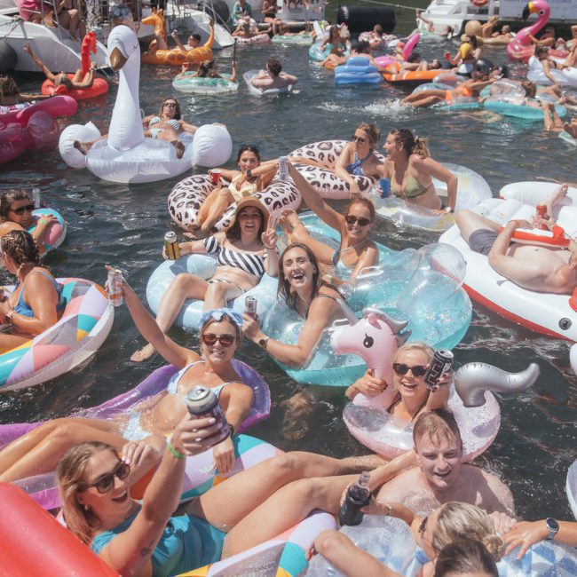 A diverse group of people on inflatable floaties, including a unicorn, donut, and cow, enjoy a sunny day on the water, holding beverages and smiling. In the background, boats and more people add to the lively atmosphere—an ideal setting for a corporate boat event in Sydney Harbour.