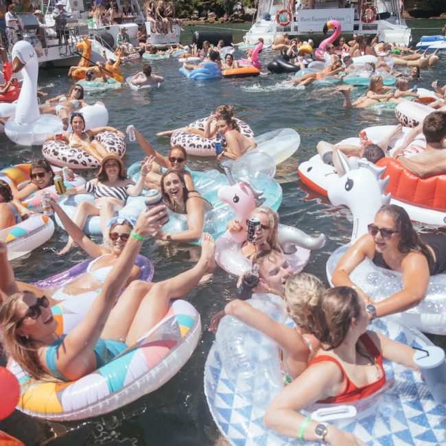A group of people on inflatable floats enjoy a sunny day on the water, surrounded by boats and other floaters. Nearby, a DJ boat hire Sydney adds lively music to the cheerful atmosphere.