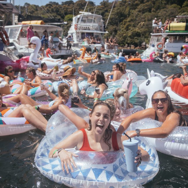 A lively scene on a lake with many people in swimsuits enjoying time on inflatable floats and boats, soaking up the sun. It's the perfect day for a Corporate boat event or even considering luxury yacht hire Sydney for a more exclusive experience.