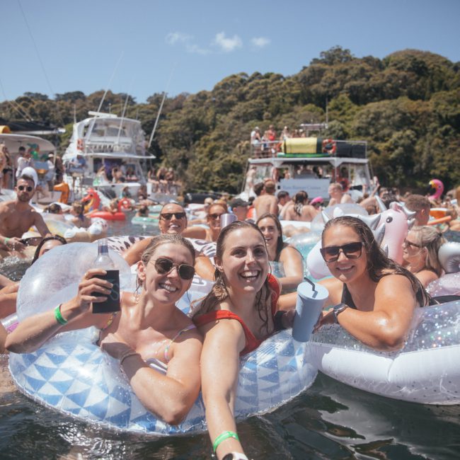 Group of people in swimsuits enjoying a sunny day on inflatable floats in the water, with dense forest in the background and multiple boats nearby, including a Catamaran party Sydney boat adding to the festive atmosphere.