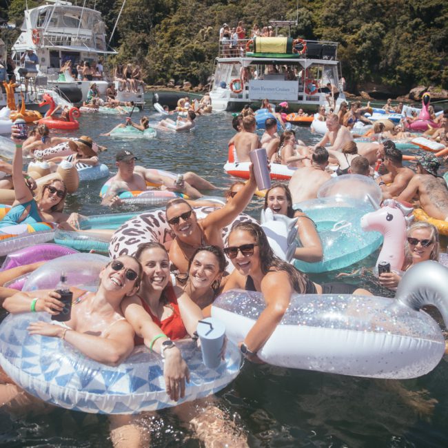A large group of people in swimsuits enjoying a lake party on various colorful inflatable floats, with boats and forested hills in the background. Consider a DJ boat hire Sydney to elevate the fun or imagine this scene during a private yacht charter Sydney Harbour for an unforgettable experience.