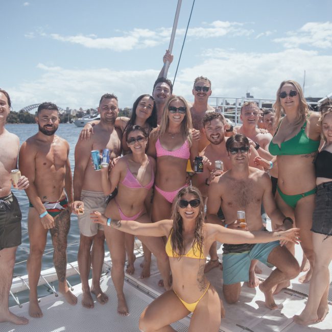 A group of twelve people in swimwear are smiling and posing on a catamaran under sunny skies, with water and the Sydney cityscape visible in the background.