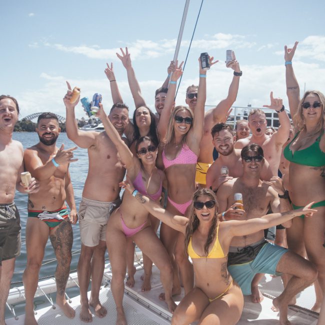 A group of people in swimwear are posing and celebrating on a catamaran party in sunny weather, with some holding drinks and raising their arms in excitement.