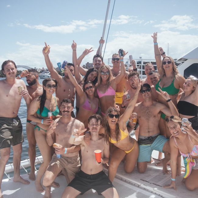A group of smiling people in swimwear posing together on a private yacht charter Sydney Harbour on a sunny day with water and cityscape in the background.
