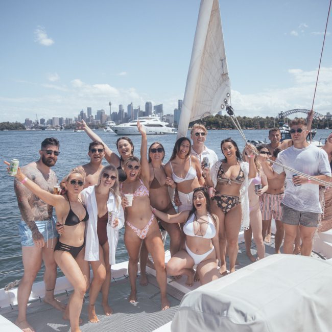 A group of people in swimwear posing together on a luxury yacht with a cityscape in the background under a clear sky.