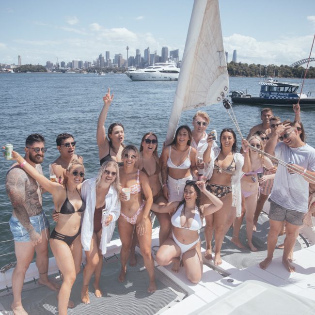 A group of people in swimwear stand on a sailboat in a harbor with a city skyline and other boats in the background. Some individuals are posing for the photo while others adjust the sail, enjoying their private yacht charter Sydney Harbour.