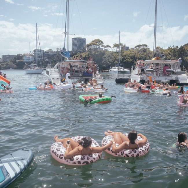 People on inflatable floats and in boats enjoy a sunny day on the water, surrounded by trees and buildings, with DJ boat hire Sydney adding vibrant music to the scene.