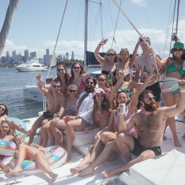 A group of people in swimwear are gathered on a boat, smiling and holding drinks, with a city skyline and other boats visible in the background. The private yacht charter Sydney Harbour provides the perfect setting for their joyful celebration.