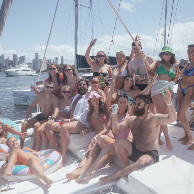 A group of people in swimwear are gathered on a luxury yacht hire in Sydney Harbour, smiling and holding drinks, against a backdrop of a city skyline and other boats.