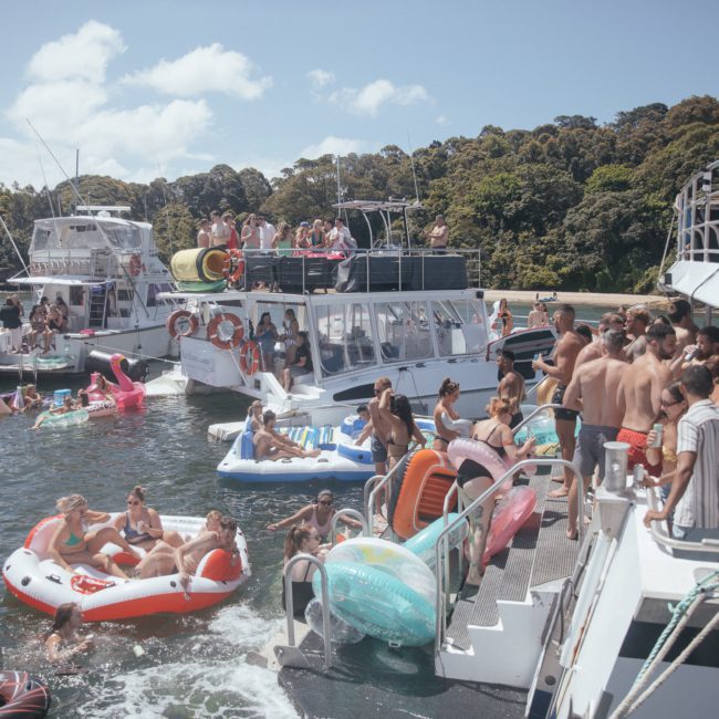 A large group of people enjoying a catamaran party near a forested shoreline, with several boats and inflatables on the water.