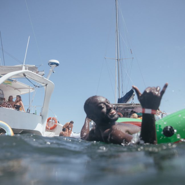 A man floats on an inflatable watermelon in the water, giving a "shaka" hand sign. Several people are enjoying themselves on a nearby sailboat, perfect for a Sydney boat party hire.
