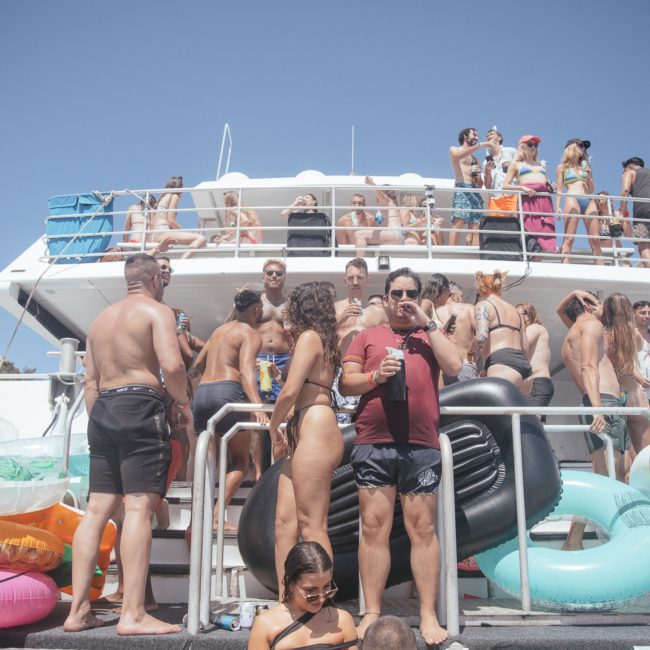 A group of people in swimwear are gathered on and around a catamaran, enjoying a sunny day. Inflatable pool toys like a flamingo and donut are visible. Trees and clear blue sky are in the background, enhancing the joyous atmosphere of this luxury yacht hire in Sydney.