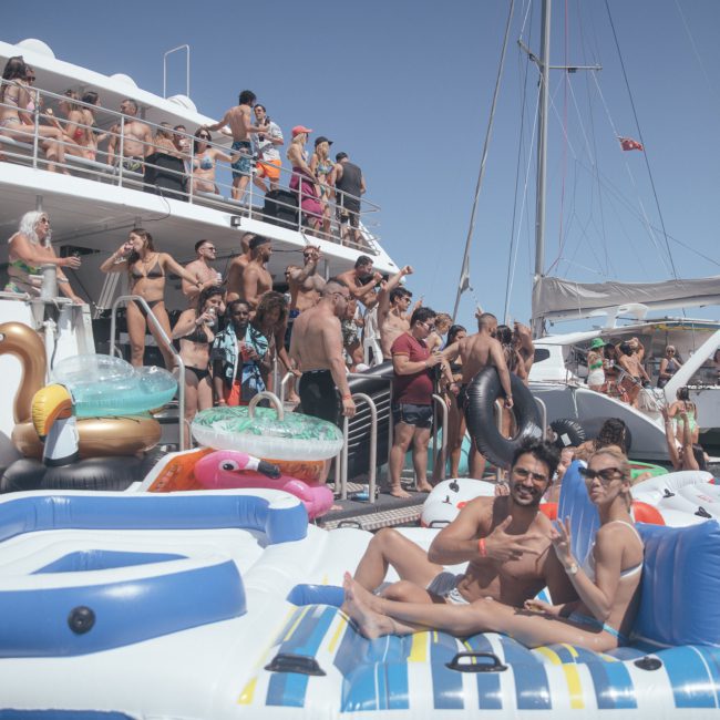 A large group of people is gathered on a boat and surrounding inflatables in the water, enjoying a sunny day. Many are in swimwear, and two men are lounging on a large inflatable raft in the foreground. Perfect for those seeking DJ boat hire Sydney, it's an ideal setting for a Catamaran party Sydney.