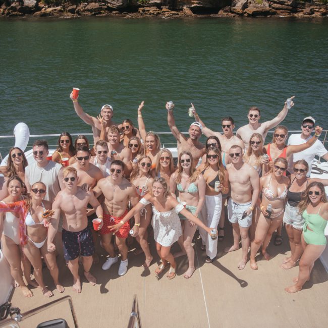 A large group of people stands and poses on the deck of a boat, many holding drinks and smiling. The boat is on green water with a rocky shoreline in the background, epitomizing the luxury yacht hire Sydney experience.
