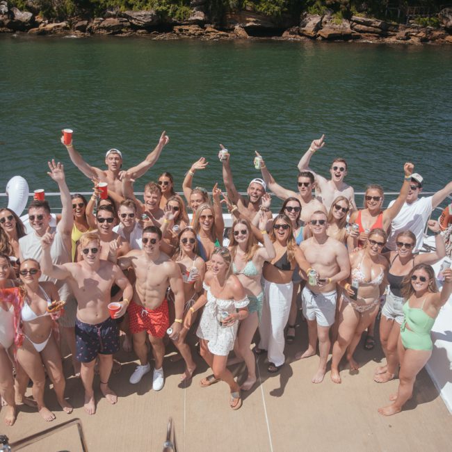 A large group of people in swimwear are enjoying a sunny day on a boat in Sydney Harbour, smiling and posing with raised hands and drinks. They are on a body of water with vegetation in the background, making it a perfect scene for a private yacht charter or corporate boat events.