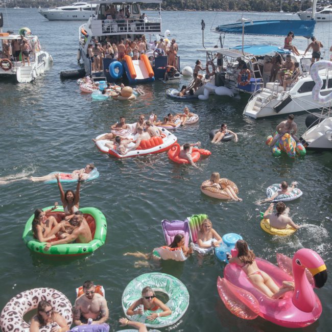 People are enjoying a sunny day on a harbor, floating on various inflatable pool toys and swimming around anchored yachts from a private yacht charter in Sydney Harbour. Many are relaxing or socializing in the water, with the city skyline visible in the background.