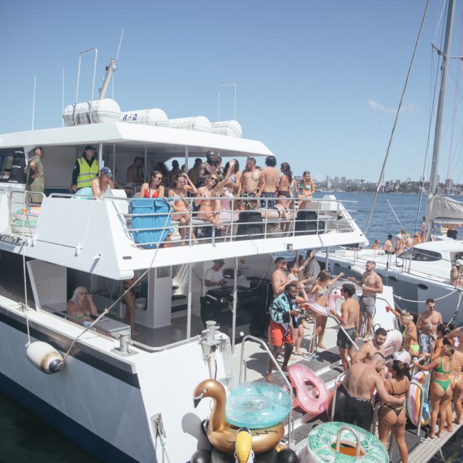 A luxury yacht hire Sydney experience with a boat crowded with people enjoying a party on the water. Inflatable pool toys, including a gold swan, are attached to the side.