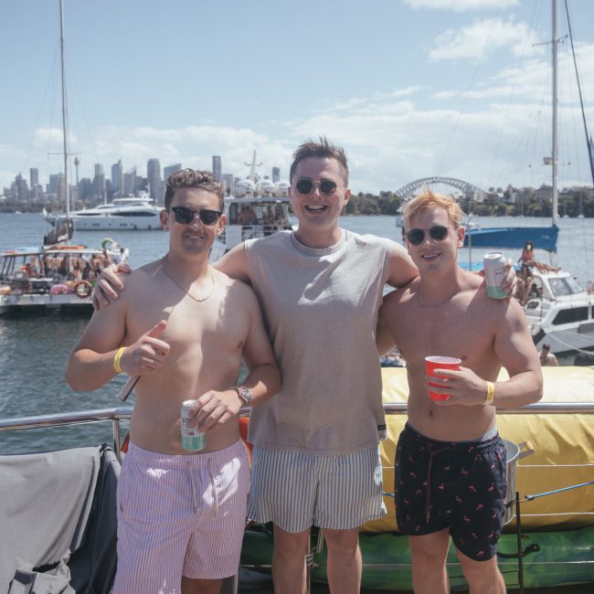 Three men in swimwear with drinks pose together on a luxury yacht, with a city skyline and other boats in the background on a sunny day.