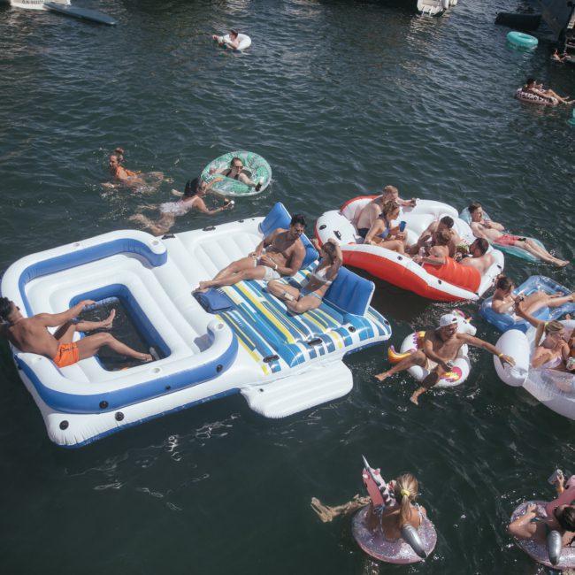 A group of people enjoy a sunny day on a lake, floating on various inflatable rafts and devices, including a large floating lounge. The water is filled with more inflatables and swimmers, reminiscent of a luxury yacht hire Sydney experience.