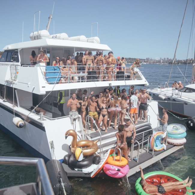 A group of people in swimwear are gathered on the deck of a luxury yacht hire in Sydney, with inflatable pool floats nearby, enjoying a sunny day on the water.