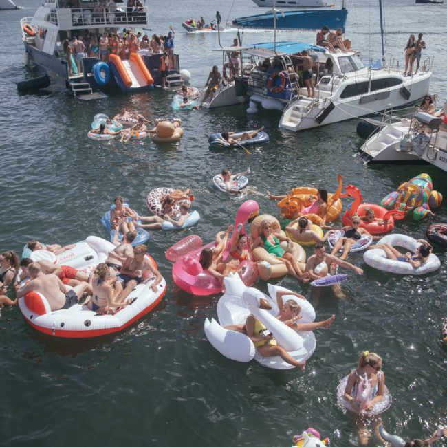 People are enjoying a sunny day on the water, floating on various colorful inflatable rafts and boats, with several larger boats anchored nearby. Some are part of a lively Sydney boat party hire, enhancing the festive atmosphere.