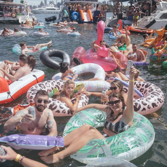 People on various inflatable floats enjoying drinks and socializing in a crowded body of water with several boats in the background, including a luxurious yacht hire Sydney that adds an extra touch of elegance to the scene.