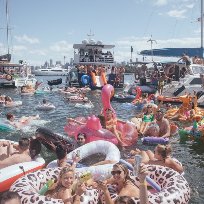 A lively scene of people on inflatable pool floats in a harbor, surrounded by boats and enjoying a sunny day. Some hold drinks, and a large inflatable water slide is visible in the background, with many considering Private yacht charter Sydney Harbour for their next adventure.