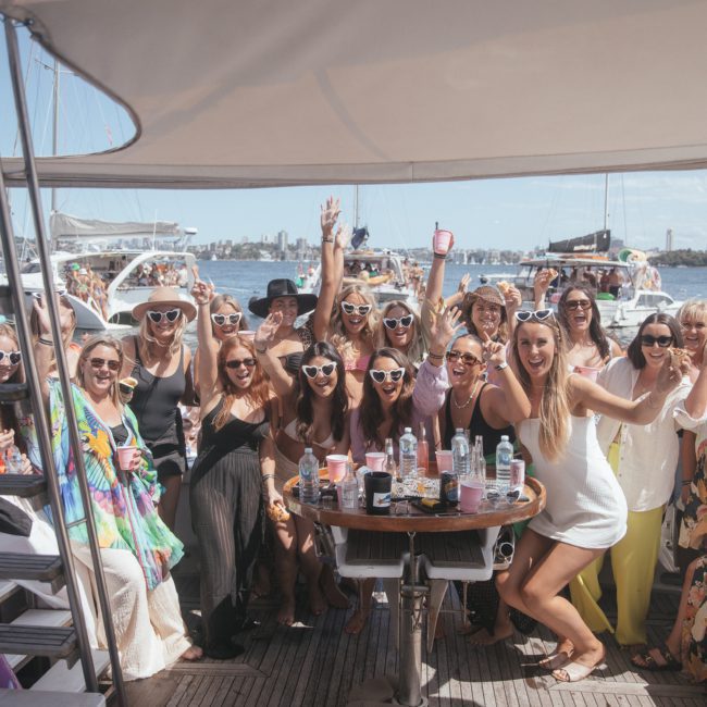 A large group of people on a boat enjoying a sunny day, many wearing sunglasses and holding drinks, with smiles and raised hands, surrounded by other boats on the water. Perfect for anyone considering a Sydney boat party hire or looking into luxury yacht hire in Sydney.
