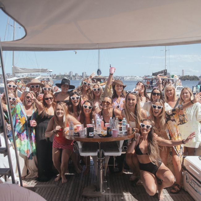 A group of people pose for a photo aboard a boat deck, wearing sunglasses and casual clothing. Various beverages and items are on a table, with other boats visible in the background on a sunny day, capturing the essence of a lively Sydney boat party hire.