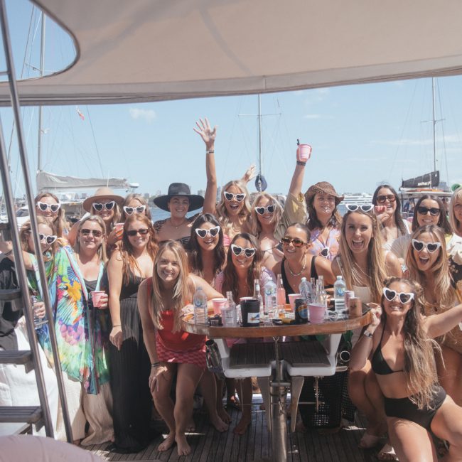 A group of people wearing heart-shaped sunglasses gather around a table on a private yacht charter in Sydney Harbour during a sunny day, posing for the camera.