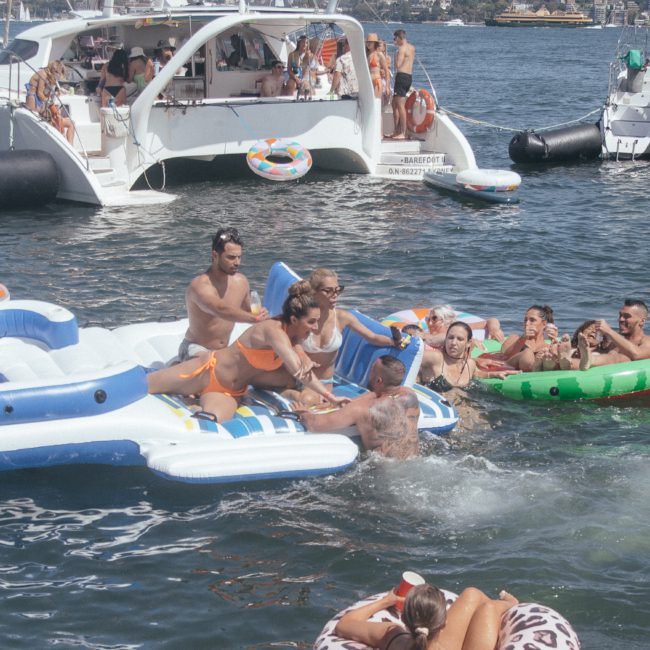 People are enjoying a sunny day on the water, floating on inflatables near boats. Some individuals are on a large inflatable platform, while others relax in smaller floaties. Consider a catamaran party Sydney-style for your next outing or opt for DJ boat hire Sydney to elevate the experience!