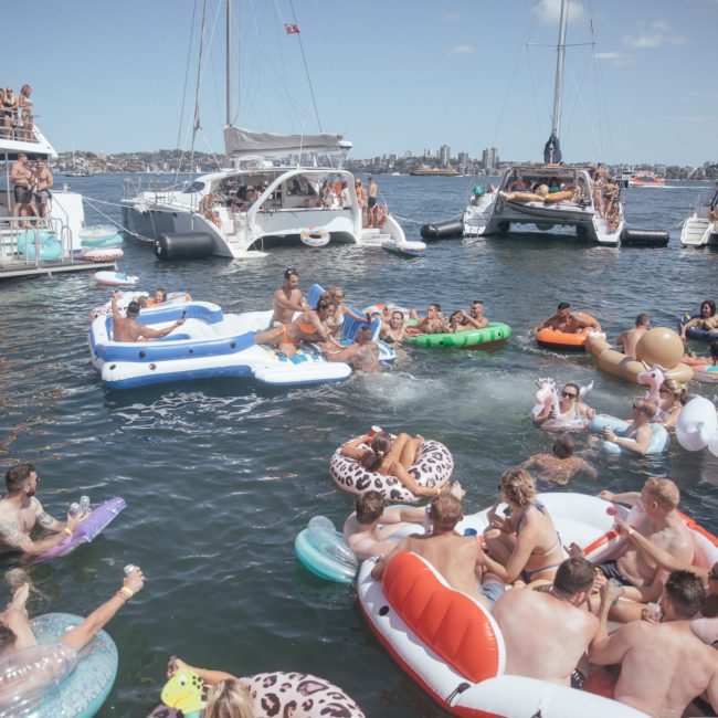 People are gathered on boats and inflatable rafts, enjoying a sunny day on the water. Several boats are anchored close together with many people swimming or floating on colorful inflatables. The scene is vibrant and lively, much like a Catamaran party in Sydney Harbour.