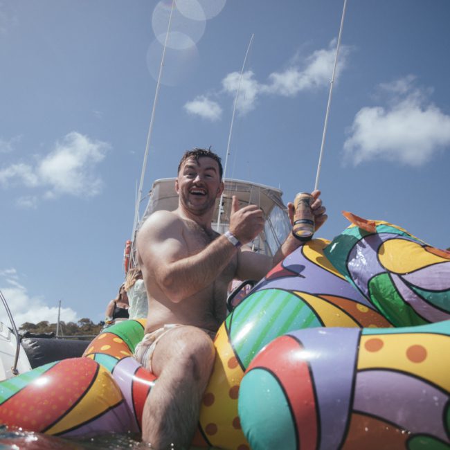 A man holding a drink is sitting on a colorful inflatable float in the water, smiling with a luxury yacht hire Sydney and clear sky in the background.