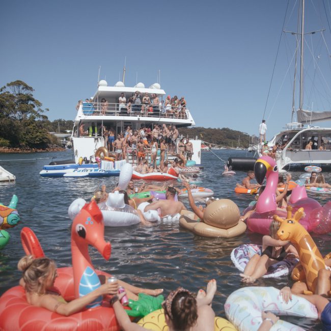 A group of people on a boat and in inflatable floaties relax in the water on a sunny day. The scene includes various inflatable animals and a background of trees and blue sky, perfect for a catamaran party Sydney style.
