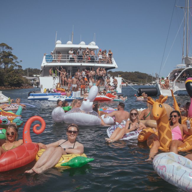 People on inflatable pool floats enjoy a sunny day in the water near boats, with a dense crowd also visible on the boats in the background, making it seem like a lively Sydney boat party hire event.