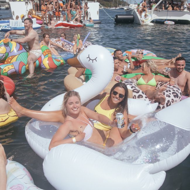 A group of people enjoying a sunny day on an array of inflatable floats in the water. Some are drinking beverages while others are mingling and smiling. Boats, including private yacht charters in Sydney Harbour, are visible in the background.
